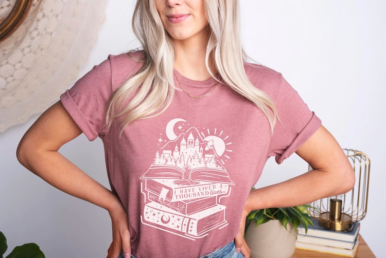 May include: A mauve t-shirt with a white graphic design of a castle and forest scene on top of a stack of books. The text on the shirt reads "I have lived a thousand lives."