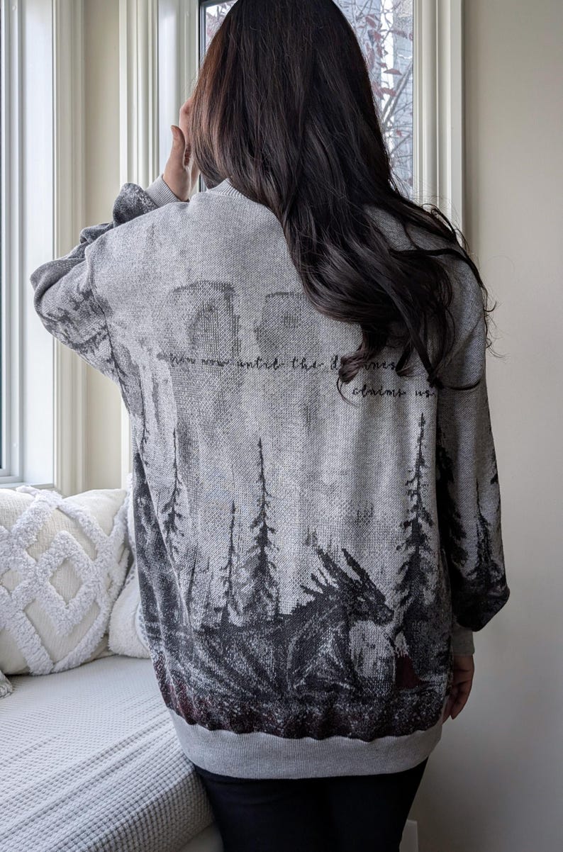 May include: Gray sweater with a forest and dragon design. The back of the sweater features a detailed illustration of a dragon, trees, and the text "Now now until the darkness claims us." The sweater is a comfortable, casual style.