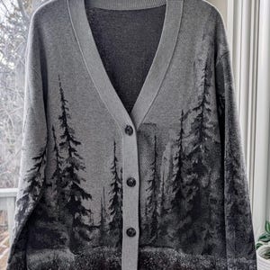 May include: A gray cardigan sweater with a V-neck and button closure. The sweater features a forest scene design in shades of black and gray. The sweater is hanging on a wooden hanger.