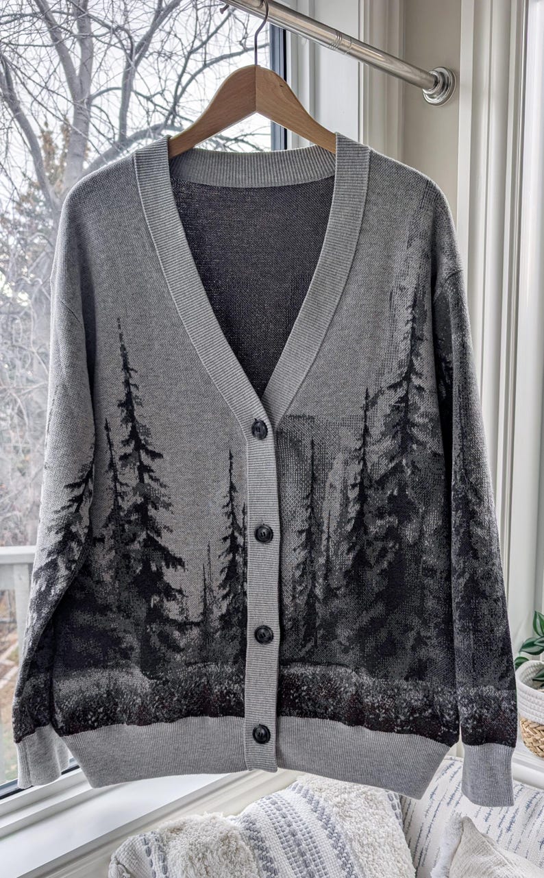 May include: A gray cardigan sweater with a V-neck and button closure. The sweater features a forest scene design in shades of black and gray. The sweater is hanging on a wooden hanger.