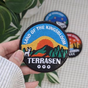 May include: Embroidered patch with a circular design featuring a mountain landscape, a deer, and the text "LAND OF THE KINGSBLOOM TERRASEN". The patch has a black border and is held in front of a plant.