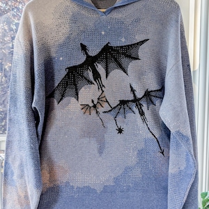 K&ouml;nnte beinhalten: Ein hellblauer Strick-Kapuzenpullover mit einer dunkelblauen und schwarzen Grafik von drei fliegenden Drachen vor einem bew&ouml;lkten Himmel. Das lang&auml;rmelige Kleidungsst&uuml;ck verf&uuml;gt &uuml;ber eine Kapuze sowie gerippte B&uuml;ndchen an &Auml;rmeln und Saum.