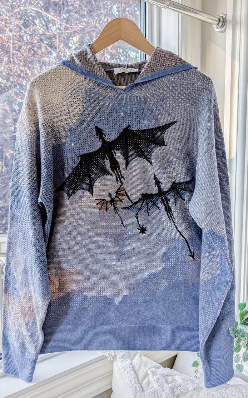 K&ouml;nnte beinhalten: Ein hellblauer Strick-Kapuzenpullover mit einer dunkelblauen und schwarzen Grafik von drei fliegenden Drachen vor einem bew&ouml;lkten Himmel. Das lang&auml;rmelige Kleidungsst&uuml;ck verf&uuml;gt &uuml;ber eine Kapuze sowie gerippte B&uuml;ndchen an &Auml;rmeln und Saum.