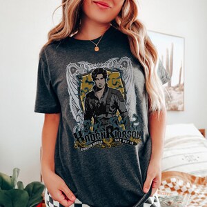 May include: A dark grey t-shirt with a graphic print of a man in a black jacket and a white shirt. The graphic also includes a shield with the text "Haden Riorsson" and "Riorson House Sat Oct 10".