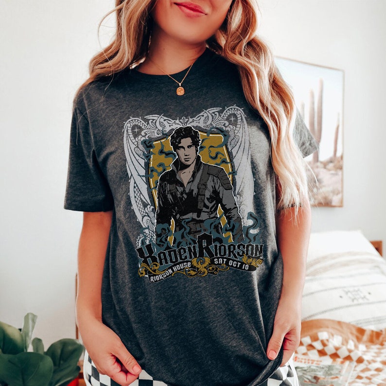May include: A dark grey t-shirt with a graphic print of a man in a black jacket and a white shirt. The graphic also includes a shield with the text "Haden Riorsson" and "Riorson House Sat Oct 10".