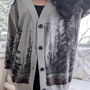 May include: Gray cardigan sweater with a forest scene print. The button-down sweater features a V-neck and long sleeves. The forest scene is printed in shades of gray and black. The sweater is worn over a black top.