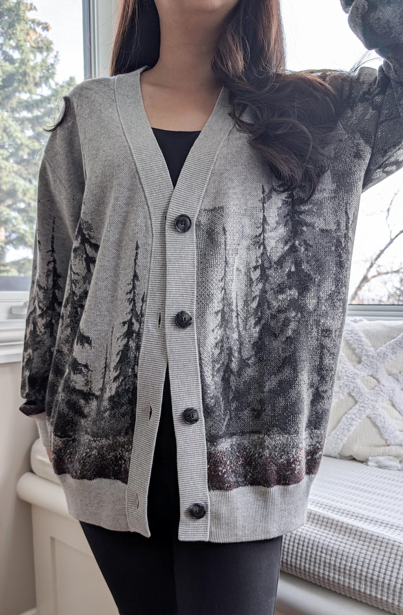 May include: Gray cardigan sweater with a forest scene print. The button-down sweater features a V-neck and long sleeves. The forest scene is printed in shades of gray and black. The sweater is worn over a black top.