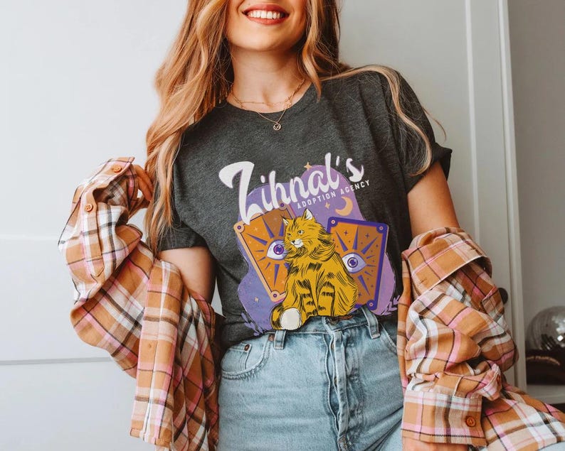 May include: A dark gray t-shirt with the text "Zihnal's Adoption Agency" and a cartoon illustration of a cat with a purple, blue, and yellow background.