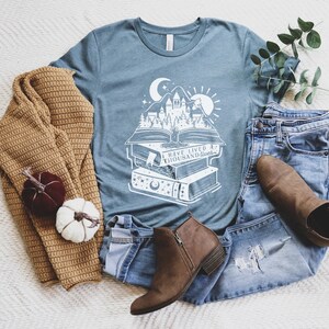 May include: A teal blue t-shirt with a white graphic of a castle rising from a stack of books. The graphic includes a crescent moon, stars, and a sun. The text on the shirt reads "I have lived a thousand lives".