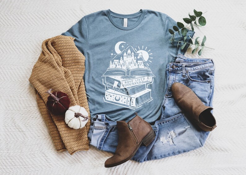 May include: A teal blue t-shirt with a white graphic of a castle rising from a stack of books. The graphic includes a crescent moon, stars, and a sun. The text on the shirt reads "I have lived a thousand lives".
