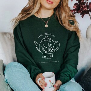 May include: A dark green sweatshirt with a white graphic of a teapot with the text "spill the tea" and "suriel tea room" below it. The text "now accepting chicken bones as payment" is at the bottom of the graphic.