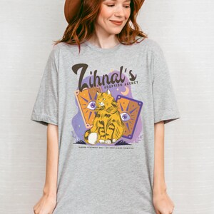 May include: A gray t-shirt with a purple and orange design featuring a cat with a yellow and orange striped pattern. The text on the shirt reads "Zihnal's Adoption Agency".