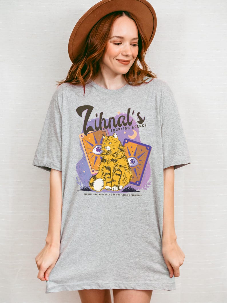 May include: A gray t-shirt with a purple and orange design featuring a cat with a yellow and orange striped pattern. The text on the shirt reads "Zihnal's Adoption Agency".