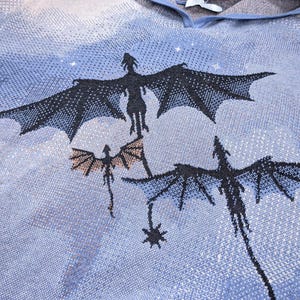 K&ouml;nnte beinhalten: Ein blauer Strick-Kapuzenpullover mit einem gewebten Motiv von drei schwarzen Drachen, die vor einem hellblauen und beigen Himmel fliegen. Der Stoff hat eine texturierte, pixelige Optik mit Kapuze und einem wei&szlig;en Innenetikett am Halsausschnitt.