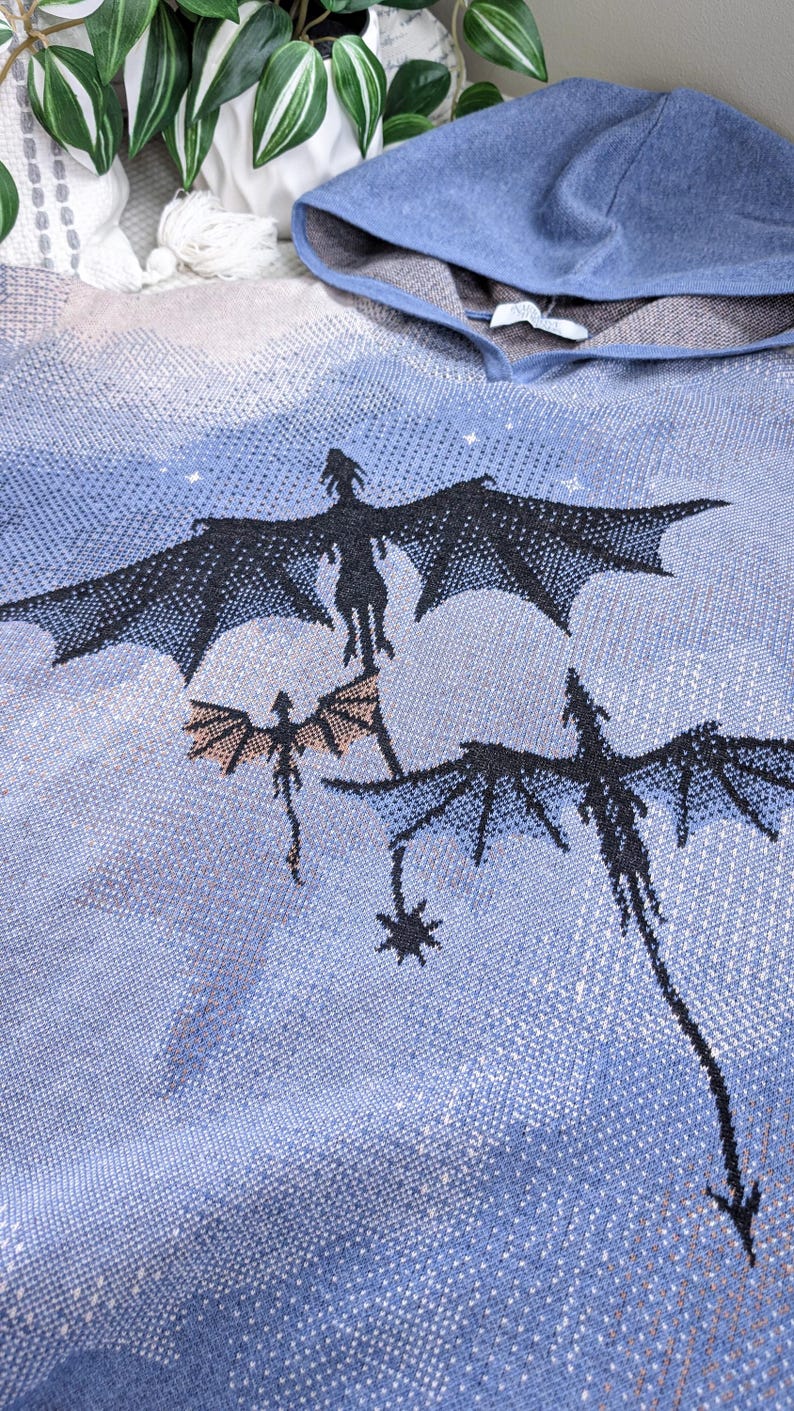 K&ouml;nnte beinhalten: Ein blauer Strick-Kapuzenpullover mit einem gewebten Motiv von drei schwarzen Drachen, die vor einem hellblauen und beigen Himmel fliegen. Der Stoff hat eine texturierte, pixelige Optik mit Kapuze und einem wei&szlig;en Innenetikett am Halsausschnitt.