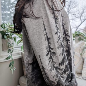 May include: A gray sweater with a forest design. The sweater features a pattern of dark gray trees against a lighter gray background. The sleeves and cuffs are a solid gray color. The sweater is made of a soft, knitted material.