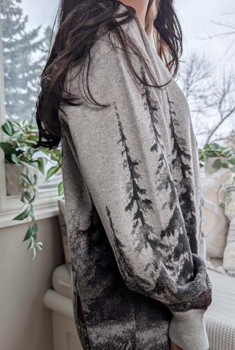 May include: A gray sweater with a forest design. The sweater features a pattern of dark gray trees against a lighter gray background. The sleeves and cuffs are a solid gray color. The sweater is made of a soft, knitted material.