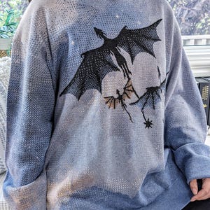 K&ouml;nnte beinhalten: Ein hellblauer Kapuzenpullover mit einem Strickmuster aus drei schwarzen fliegenden Drachen vor einem Hintergrund aus dezenten wei&szlig;en und beigen Wolken. Das Kleidungsst&uuml;ck hat lange &Auml;rmel, einen gerippten Saum und eine Kapuze mit Grafikdesign.