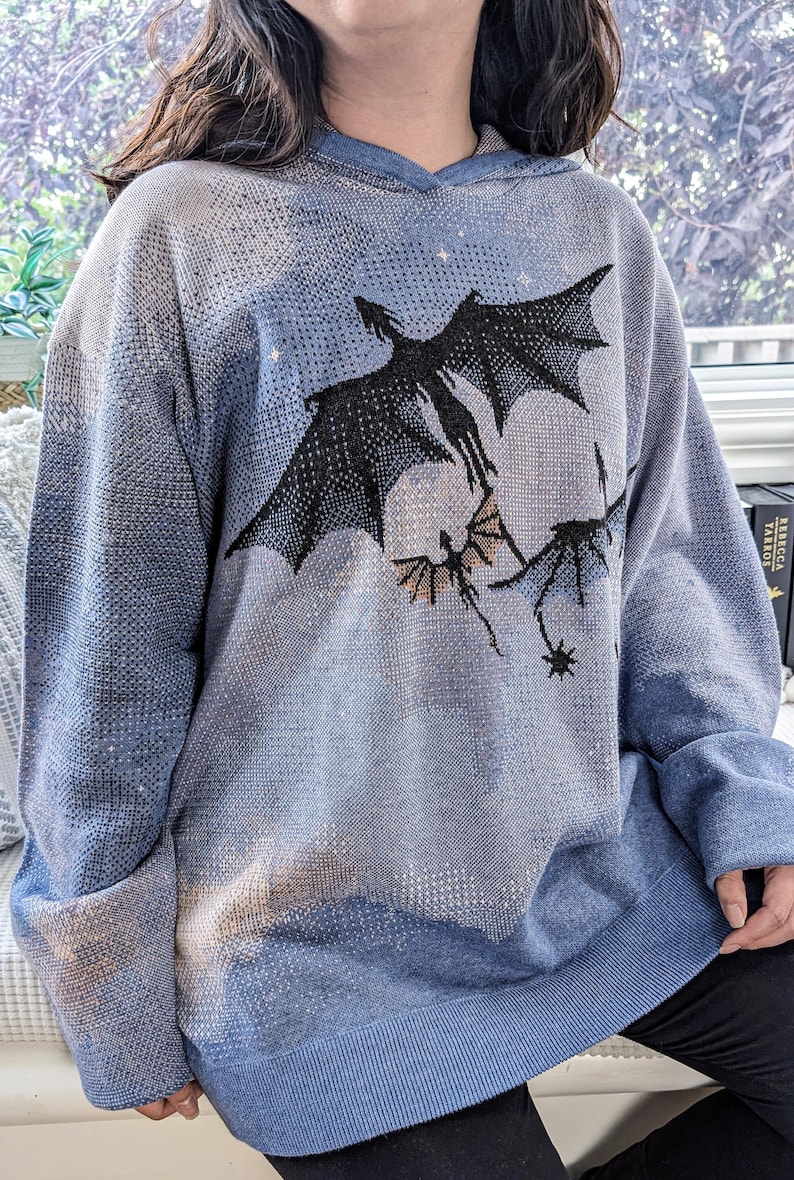 K&ouml;nnte beinhalten: Ein hellblauer Kapuzenpullover mit einem Strickmuster aus drei schwarzen fliegenden Drachen vor einem Hintergrund aus dezenten wei&szlig;en und beigen Wolken. Das Kleidungsst&uuml;ck hat lange &Auml;rmel, einen gerippten Saum und eine Kapuze mit Grafikdesign.