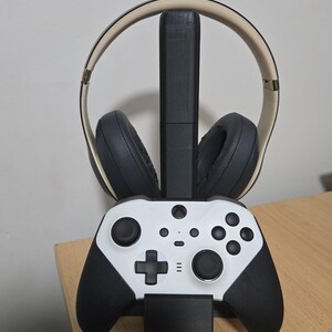 May include: A black and white gaming controller and a black headset with a beige headband are on a black stand. The stand is on a light brown wooden surface.