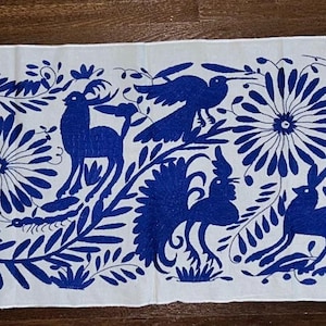 Peut inclure: Une longue pièce de tissu rectangulaire blanche avec un motif brodé bleu vif. Le motif comprend des fleurs stylisées, des oiseaux et des cerfs, créant un motif inspiré de la nature. La broderie est détaillée et couvre toute la surface.