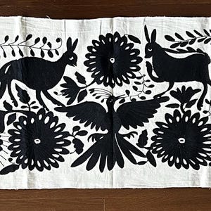 May include: A long, rectangular white table runner with a repeating black embroidered pattern of stylized animals, birds, flowers, and foliage. The runner is made of a natural fabric and is suitable for a dining table or sideboard.