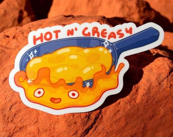 May include: A white sticker with a cartoon illustration of a frying pan with a yellow egg inside. The egg is on fire and has a happy face. The text "HOT N' GREASY" is above the pan.