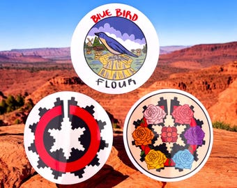 May include: Three round stickers with different designs. The first sticker has a blue bird on a pile of wheat with the text "Blue Bird Flour". The second sticker has a red and black geometric design. The third sticker has a floral design with seven colorful roses.