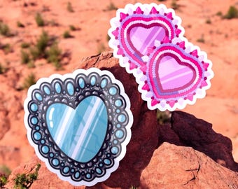 May include: Two pink heart-shaped stickers with intricate designs, and one blue heart-shaped sticker with a black and white patterned border. The stickers are on a red rock surface.