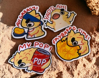 May include: Four stickers featuring cartoon bearded dragons with different expressions and text. The stickers are on a brown sandy surface. The text on the stickers reads "All Greasy!", "Yadillah!", "Frybread Time!" and "My Pop!"
