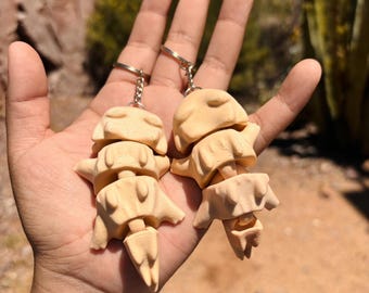 May include: Two beige clay keychain charms shaped like stacked figures with closed eyes. The charms are attached to silver key rings.