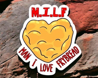 May include: A sticker with a heart-shaped frybread design. The text "M.I.L.F" is at the top in red, and "MAN I LOVE FRYBREAD" is around the heart in red. The sticker is on a textured, reddish-brown surface.