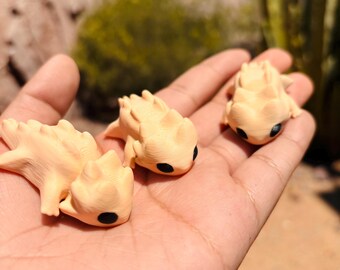 May include: Three small, beige, ceramic figurines of spiky, lizard-like creatures with black eyes. The figurines are sitting in the palm of a hand.