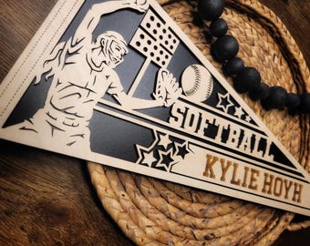 May include: A wooden softball pennant with a black background and a white silhouette of a player catching a ball. The pennant has the text "Softball" and "Kylie Hoyh" in gold lettering.