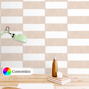 Op de afbeelding: Een beige en wit gestreept behang met een bureau op de voorgrond. Het bureau heeft een houten oppervlak en een lamp, een waterfles, een kaars en een stapel notitieboekjes erop. Het behang heeft een "Aanpassen"-knop met een regenboogkleurenwiel.