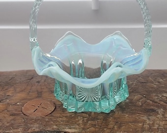 May include: A small, light blue glass basket with a ruffled edge and a woven handle. The basket is made of iridescent glass and has a delicate, feminine look.