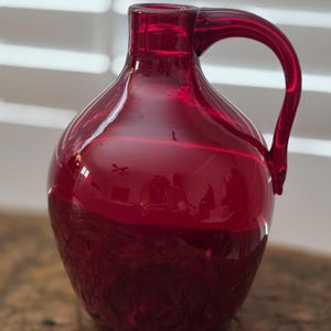 May include: A ruby red glass pitcher with a rounded body and a curved handle. The pitcher sits on a wooden surface, reflecting light and shadows. The handle is attached near the top of the pitcher, and the opening is visible.