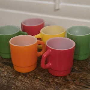 May include: A collection of six stackable mugs in vibrant colors including orange, red, yellow, and two shades of green. Each mug features a smooth, glossy finish, a rounded handle, and a distinct stepped base design for stacking functionality.