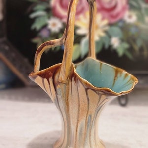 May include: A small ceramic vase with a handle in the shape of a basket. The vase is decorated with brown, blue, and yellow glaze, creating a marbled effect.