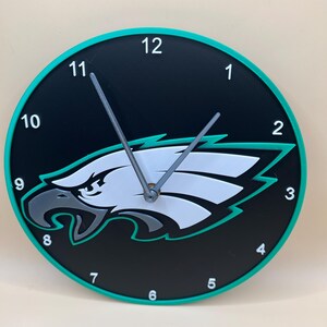 Könnte beinhalten: Eine schwarz-weiße Uhr mit grünem Rand. Das Zifferblatt der Uhr zeigt einen stilisierten Adlerkopf mit einem weißen Flügel und einer grünen Umrandung. Die Zahlen 1 bis 12 sind in Weiß um den Rand des Zifferblatts gedruckt.