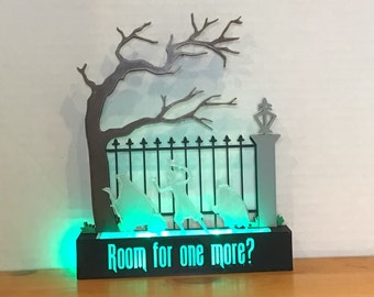 May include: A black wooden base with a green light and a white silhouette of a gate, a tree, and three figures. The base has the text "Room for one more?" printed on it.