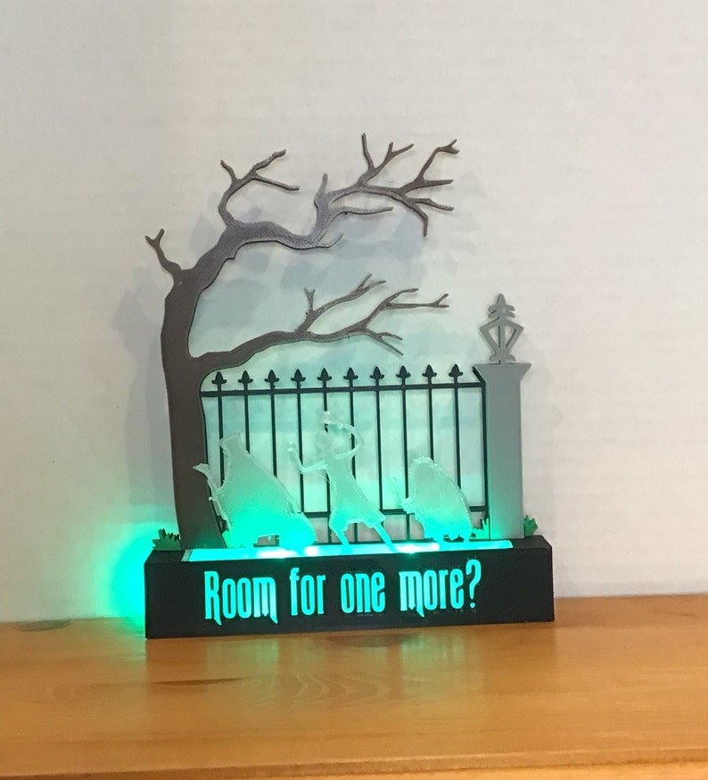 May include: A black wooden base with a green light and a white silhouette of a gate, a tree, and three figures. The base has the text "Room for one more?" printed on it.