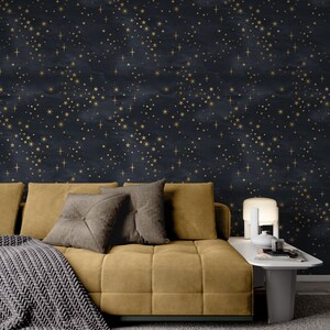 K&ouml;nnte beinhalten: Ein senfgelbes Sofa mit grauen Kissen und einer grauen Strickdecke. Das Sofa steht vor einer Wand mit einem dunkelblauen und goldenen Sternenmuster.