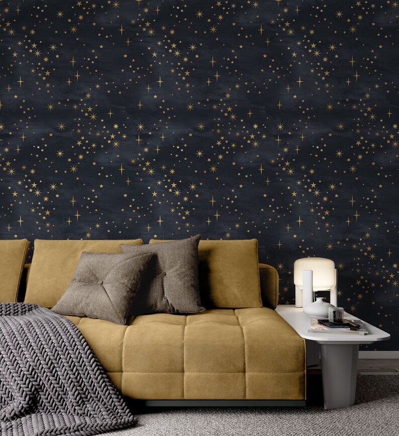 K&ouml;nnte beinhalten: Ein senfgelbes Sofa mit grauen Kissen und einer grauen Strickdecke. Das Sofa steht vor einer Wand mit einem dunkelblauen und goldenen Sternenmuster.