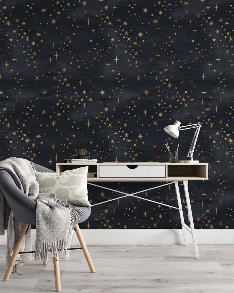 K&ouml;nnte beinhalten: Ein wei&szlig;er Schreibtisch mit einer Schublade und einer wei&szlig;en Lampe steht vor einer dunkelblauen Wand mit einem goldenen Sternenmuster. Ein grauer Sessel mit einer wei&szlig;en Wolldecke steht im Vordergrund.