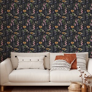 May include: A white couch with two throw pillows and a patterned throw blanket sits in front of a wall covered in a dark floral wallpaper. The wallpaper features a repeating pattern of pink, yellow, and blue flowers on a dark background.