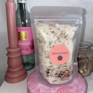 May include: A clear plastic bag filled with bath salts, labeled "Coven of Genesis" with a pink sticker. The salts are mixed with dried herbs and flower petals. A pink candle and a bottle of Moscato wine are in the background.