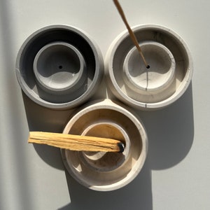 Könnte beinhalten: Drei runde Beton-Räucherstäbchenhalter in Grau, Weiß und Creme. Ein Holz-Räucherstäbchen brennt im cremefarbenen Halter.