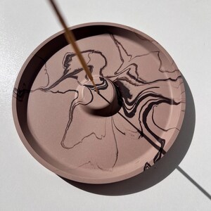 May include: A round, dusty rose incense holder with a dark brown, marbled design. The holder has a small hole in the center to hold an incense stick. The incense holder is on a white surface.