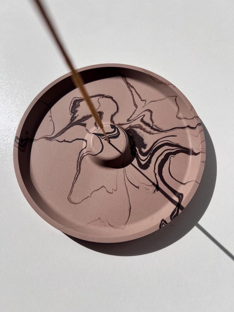 May include: A round, dusty rose incense holder with a dark brown, marbled design. The holder has a small hole in the center to hold an incense stick. The incense holder is on a white surface.